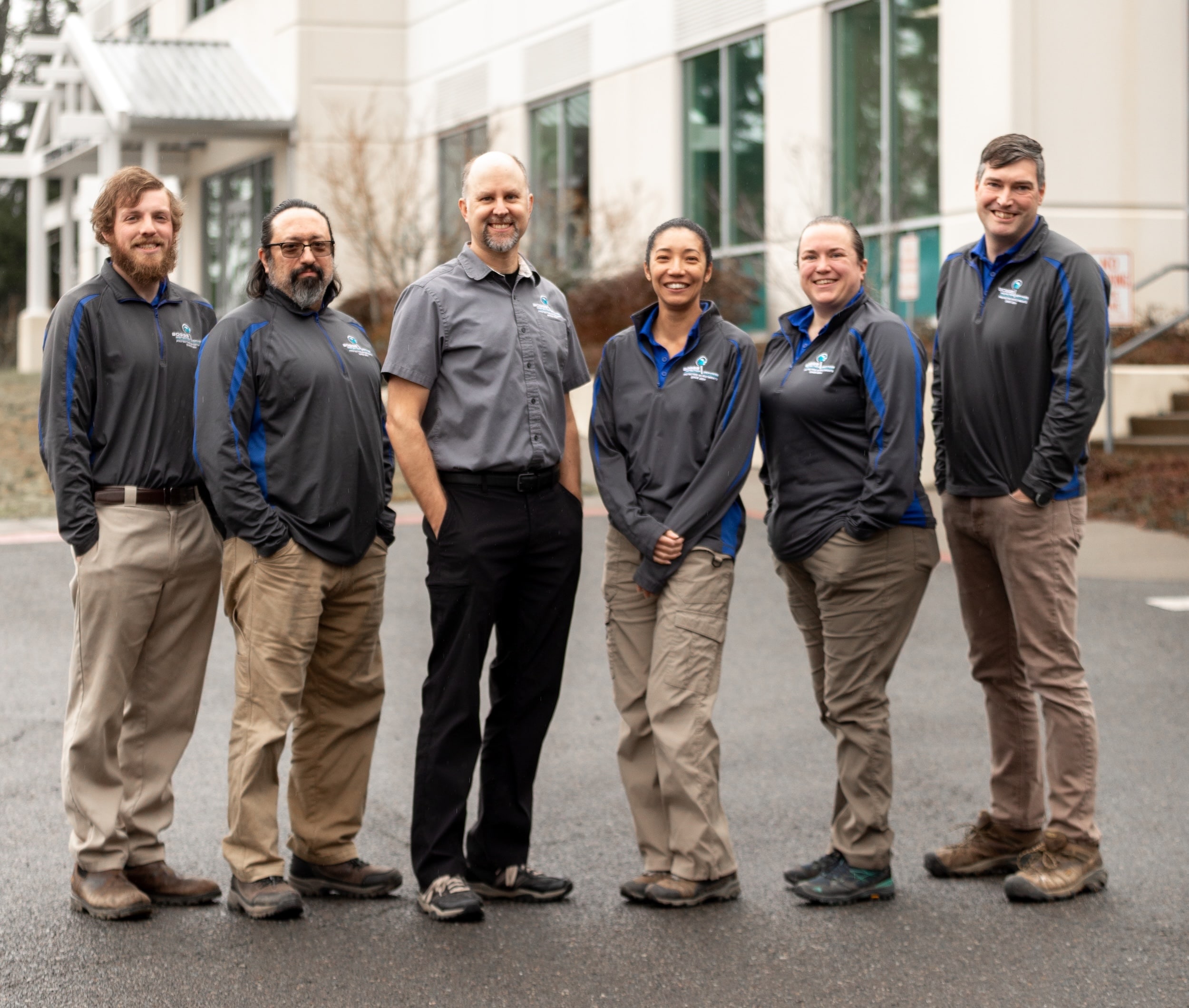 Group photo of six Boggs Inspection Services team members standing together outdoors in front of a commercial building. All wear matching dark gray and blue company polo shirts with the Boggs Inspection Services logo. The team includes four men and two women, dressed in business-casual attire with khaki or dark pants and work boots, smiling at the camera.