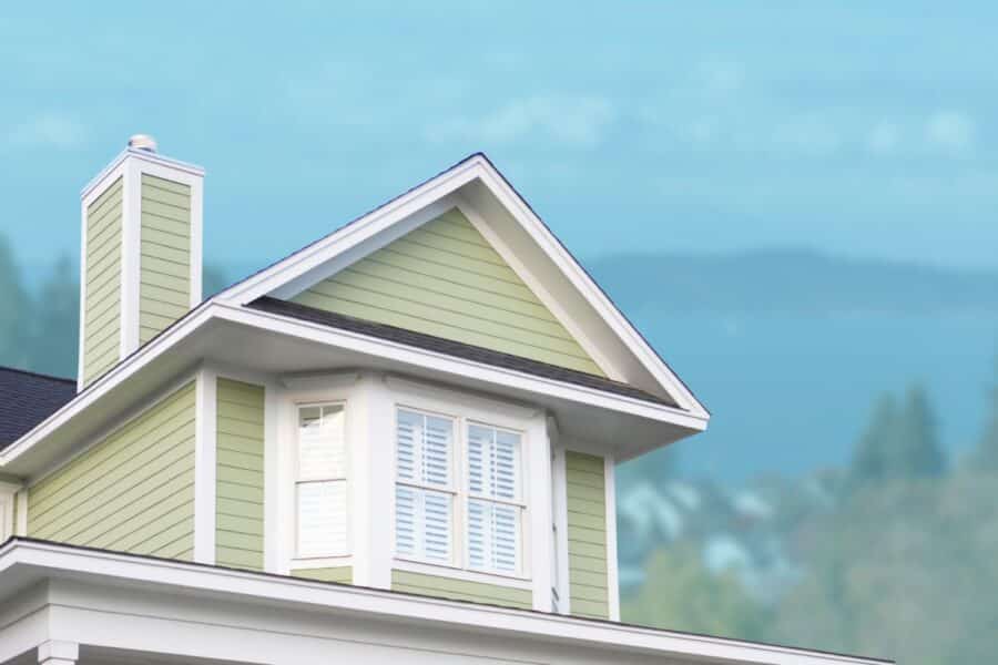 The upper exterior of a light green house with white trim and a chimney against a blurred background of trees and mountains.
