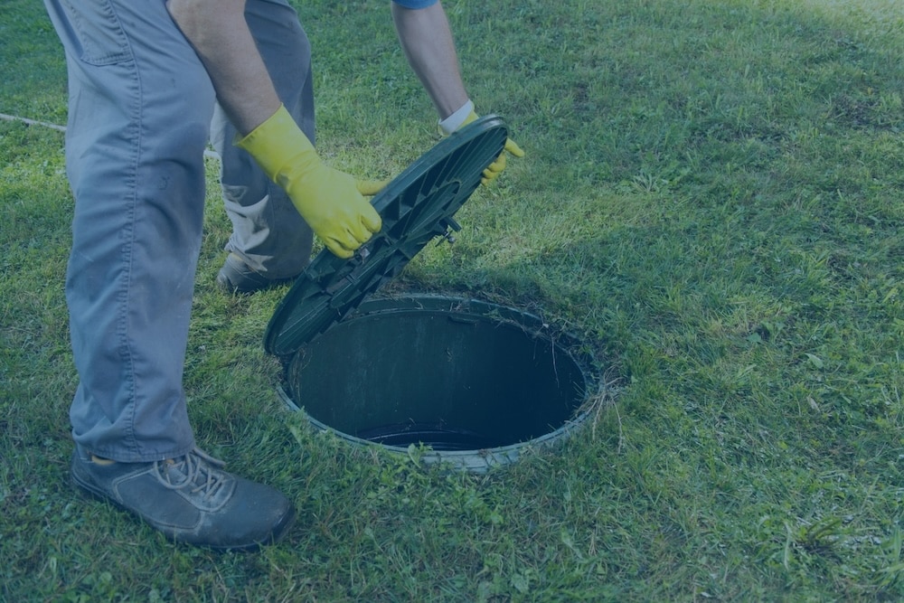 A technician wearing yellow gloves lifts the green lid of an underground septic tank riser in a grassy lawn.