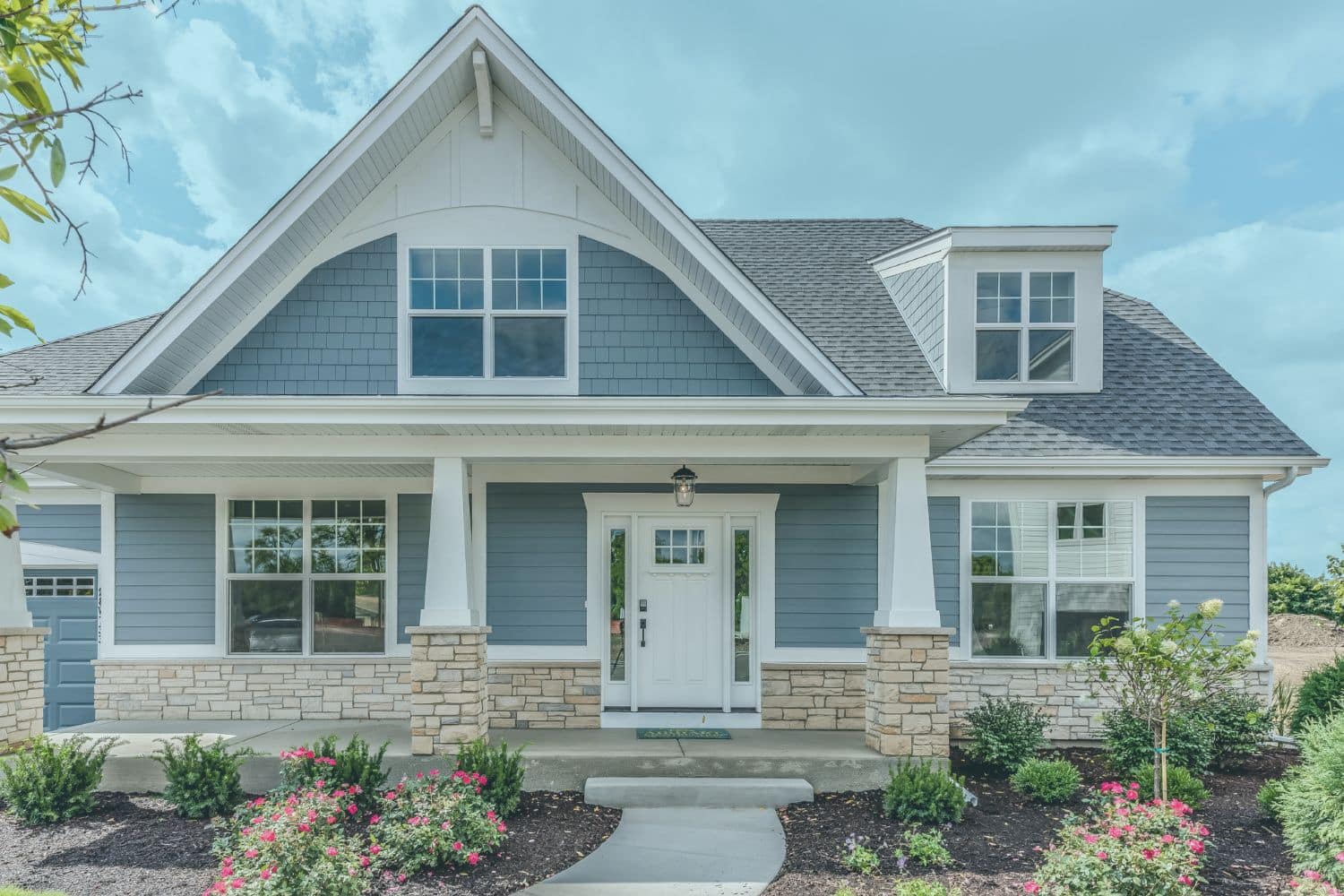 A charming blue Craftsman-style home featuring a spacious front porch with stone-based columns and neatly manicured landscaping.